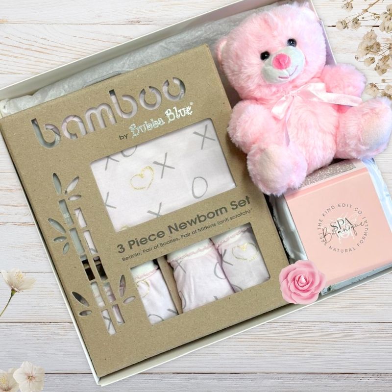 Bamboo by Bubba Blue baby gift set with pink teddy bear, clothing, and spa product in gift box on a wooden surface.
