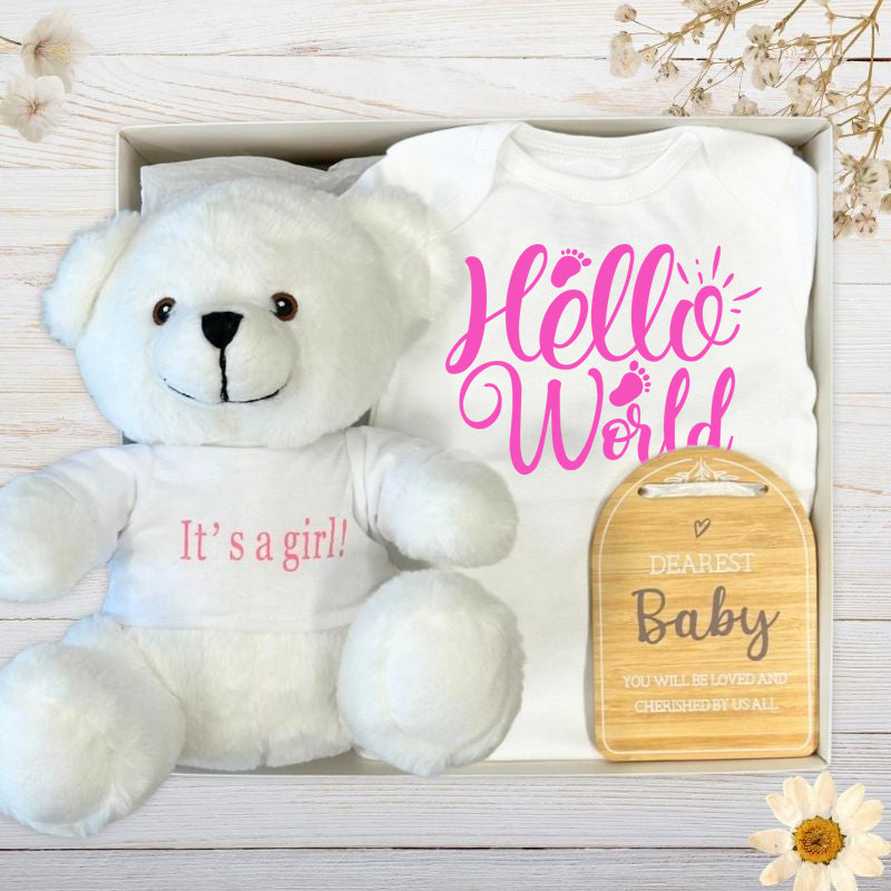 Baby gift set with white its a girl teddy bear, baby clothes, and wooden plaque on a wooden surface