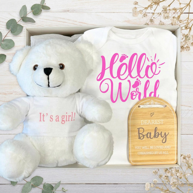Baby gift set with white its a girl teddy bear, baby clothes, and wooden plaque on a wooden surface