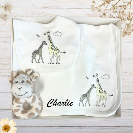 white baby blanket and bib set with giraffe prints and rattle in a box on a wooden surface with greenery.