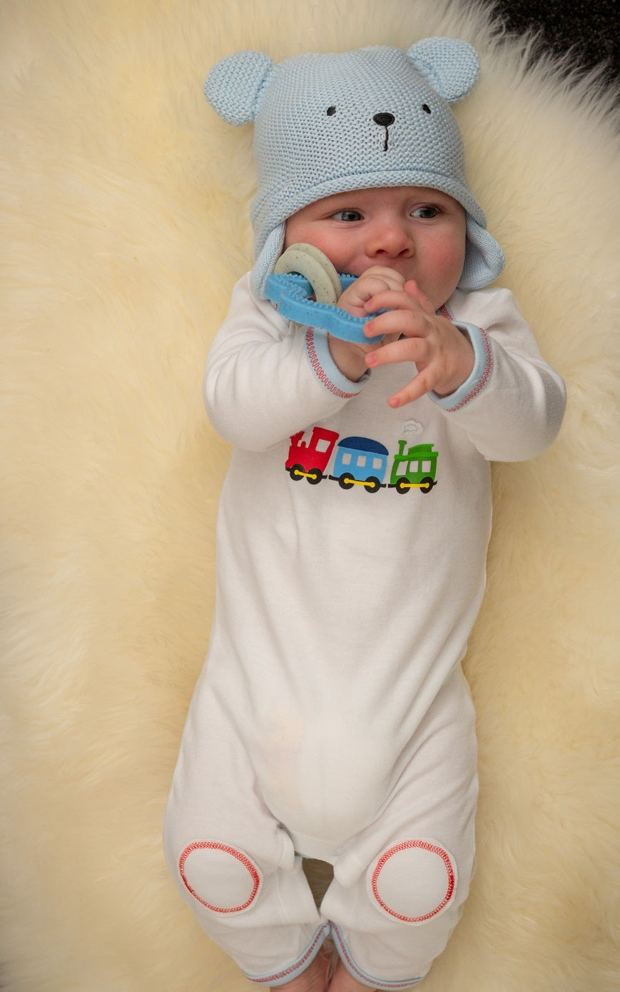 Baby in a train-themed outfit with a blue hat and colorful train design on a soft surface