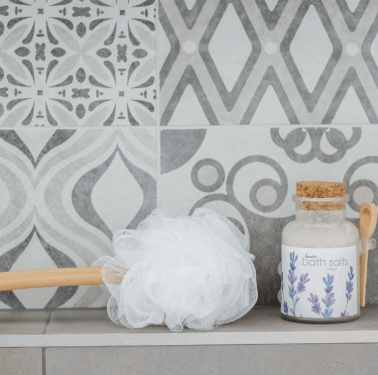Bath salt jar with lavender design and a loofah on a tiled bathroom shelf.