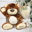 Brown teddy bear with 'Max' on its paw next to a bouquet of white flowers on a light wooden surface.