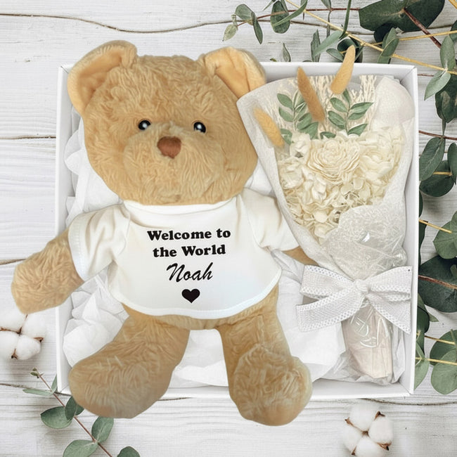 Brown teddy bear with Welcome to the Word 'Noah' on its chest next to a bouquet of white flowers on a light wooden surface.
