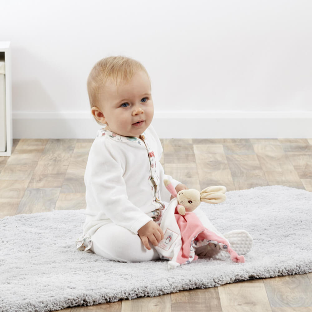 Baby sitting on a rug holding a plush Beatrix Potter Flopsy toy in a room with a light-colored floor.