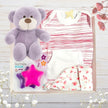 Baby gift set with red stripe onesie, purple teddy bear, floral hat, and star-shaped teether on a wooden surface.