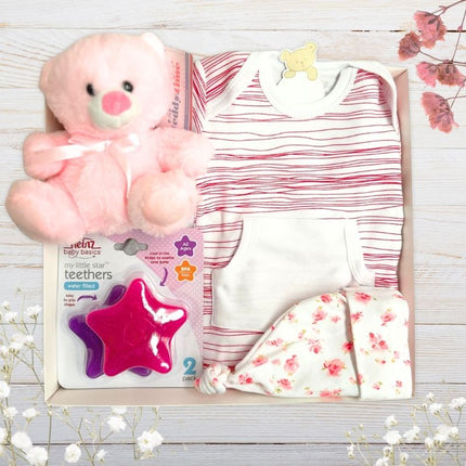 Baby gift set with red stripe onesie, pink teddy bear, floral hat, and star-shaped teether on a wooden surface.