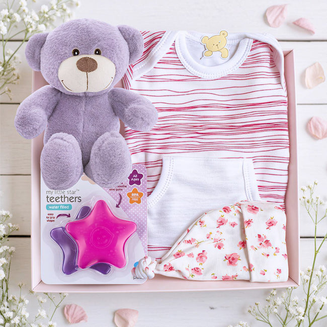 Baby gift set with red stripe onesie, purple teddy bear, floral hat, and star-shaped teether on a wooden surface.
