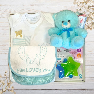 Baby gift set with teddy bear, clothes, and toys on a wooden surface