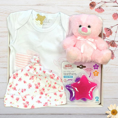 Baby gift set with white onesie, pink teddy bear, floral hat, and star-shaped teether on a wooden surface.