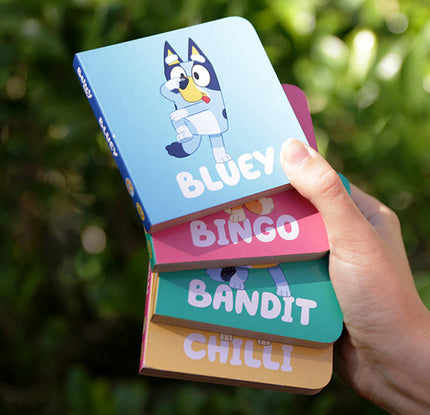 Stack of colorful books with Bluey characters held by a hand against a green leafy background