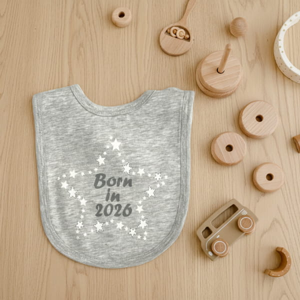 Grey bib with 'Born in 2026' text and wooden teething rings on a light  background