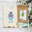 Baby romper with robot design and 'Jack' text, baby rattle, and gift box on a wooden surface.