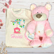 Baby outfit with embroidered design and pink teddy bear on a wooden surface.