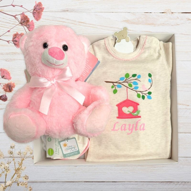 Baby outfit with embroidered design and pink teddy bear on a wooden surface.