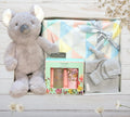 Gray plush koala toy, baby blanket, and bath products on a wooden surface with greenery.