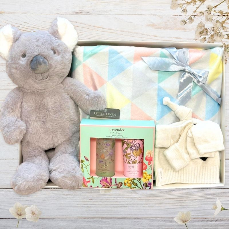 Gray plush koala toy, baby blanket, and bath products on a wooden surface with greenery.