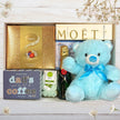 Gift set with Lindor chocolate, Moét champagne, and a blue teddy bear on a wooden surface.