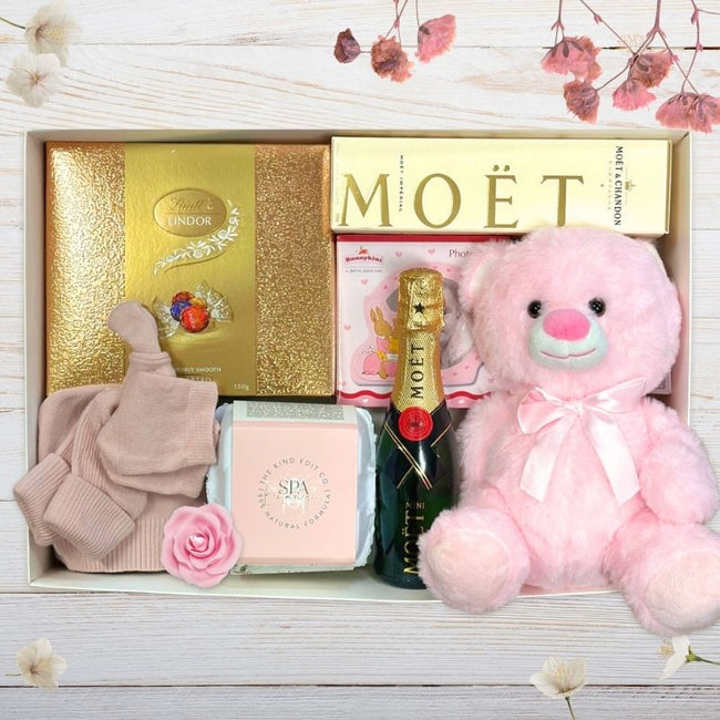 Gift box with Moét champagne, pink teddy bear, and other baby girl gift items on a wooden surface.