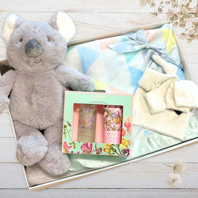 Baby gift set with plush koala, baby blanket, and skincare products on a wooden surface.