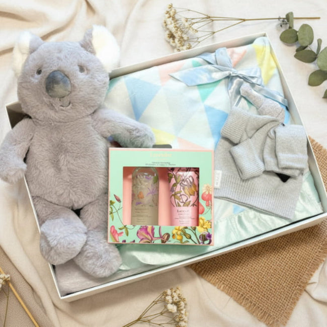Baby gift set with plush koala, baby blanket, and skincare products on a natural surface.