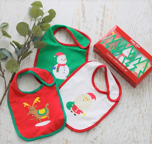 Three Christmas-themed baby bibs on a wooden surface with a box of additional bibs.