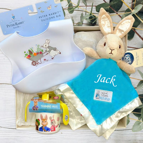 Peter Rabbit-themed baby product set including a silicone bib, training mug, and comfort blanket on a light wooden surface.