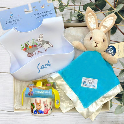 Peter Rabbit-themed baby product set including a silicone bib, training mug, and comfort blanket on a light wooden surface.