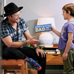 Man wearing a black hat and plaid shirt sitting on a stool, child giving him a 'Congratulations Dad' box of socks.