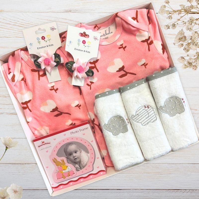 Baby gift set with pink outfit, photo frame, and elephant-themed blankets on a wooden surface.