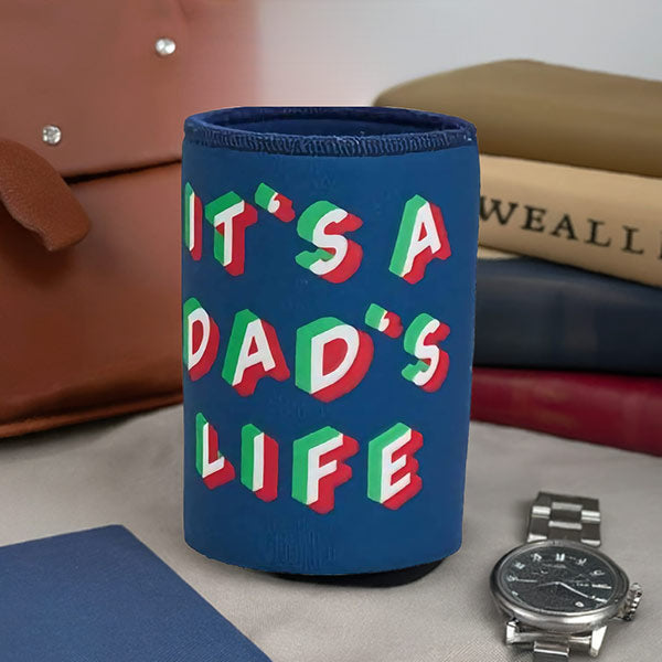 Blue can cooler with 'It's a Dad's Life' text on a surface with books and a watch.