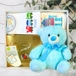 Baby gift set with blue teddy bear, baby clothes, and products on a wooden surface.