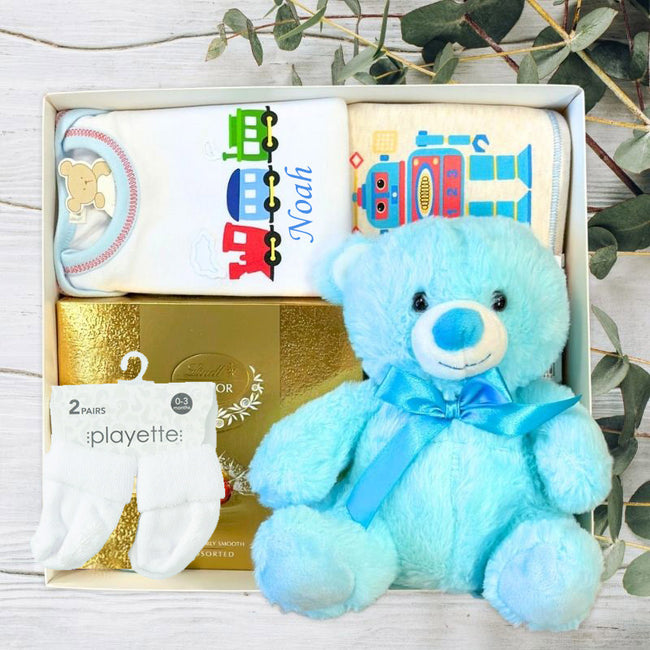 Baby gift set with blue teddy bear, baby clothes, and products on a wooden surface.