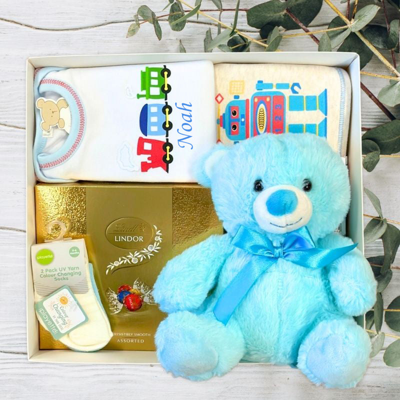Baby gift set with blue teddy bear, baby clothes, and products on a wooden surface.