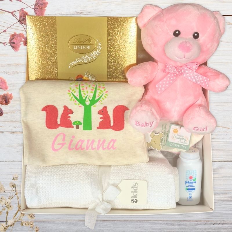 Baby gift set with pink teddy bear, blanket, and lotion bottle in a gift box on a wooden surface.