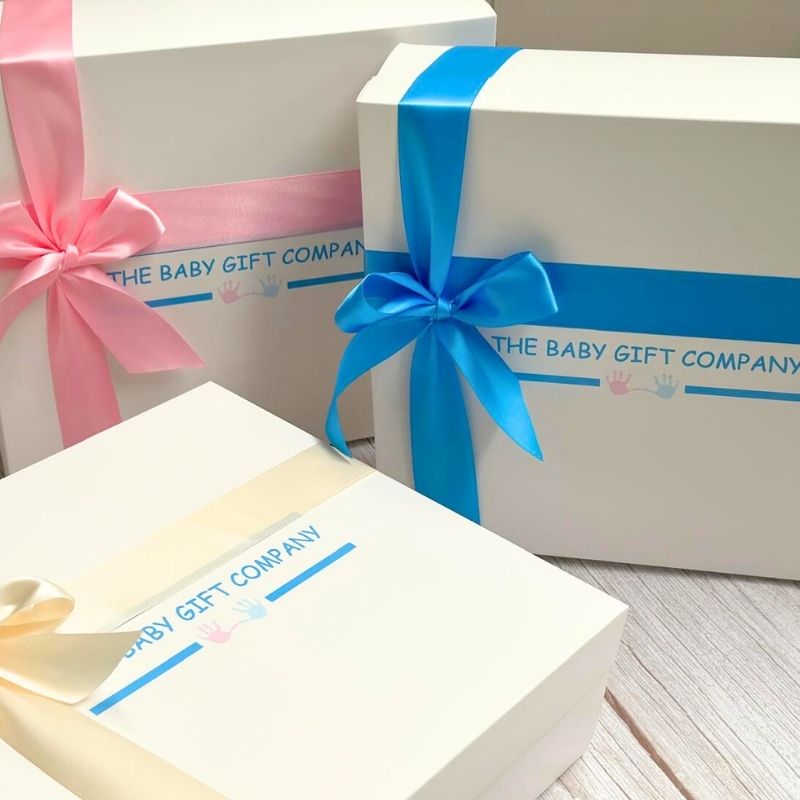 Three gift boxes from The Baby Gift Company with colorful ribbons on a light wooden surface.