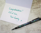 Greeting card with 'Congratulations! Lots of love, Your family x' text and a decorative pen on a textured surface.