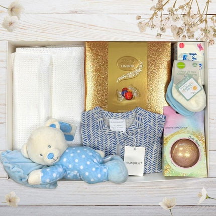 Baby gift set with teddy bear, Lindor chocolate box, and other items on a wooden surface.