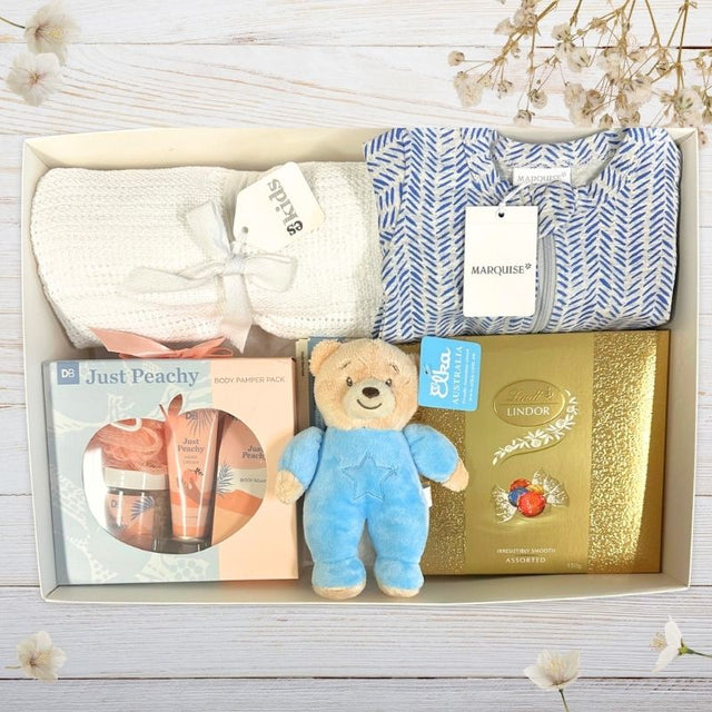 Baby gift set with teddy bear, Lindor chocolate box, and other items on a wooden surface.