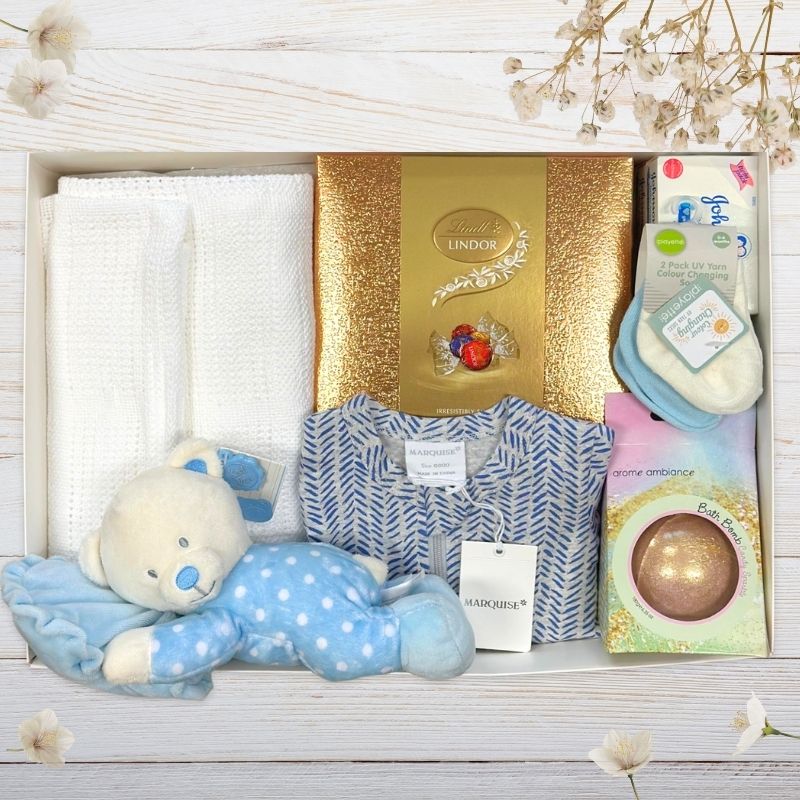 Baby gift set with teddy bear, Lindor chocolate box, and other items on a wooden surface.