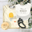 Baby gift set with romper, bib, bootie, and hello world plaque on a wooden surface with greenery.