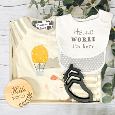 Baby gift set with romper, bib, bootie, and hello world plaque on a wooden surface with greenery.