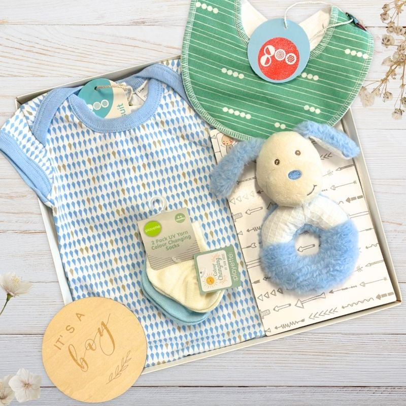 Baby boy gift set with onesie, bib, rattle, and burp cloth on a wooden surface.