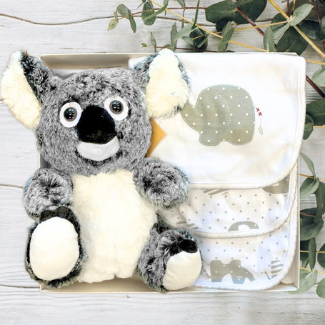 Plush koala toy with a baby clothing set in a gift box on a wooden surface with eucalyptus leaves.
