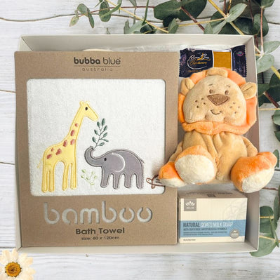 Baby gift set with lion plush toy, and baby bath towel with jungle animals, plus soap and chocolate on a wooden surface with greenery.