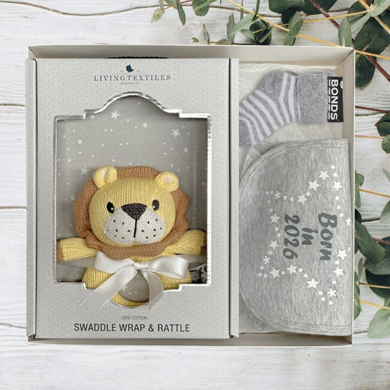 Baby gift set with lion swaddle wrap and rattle, and baby blanket with 'Born in 2026' text, on a wooden surface with greenery.