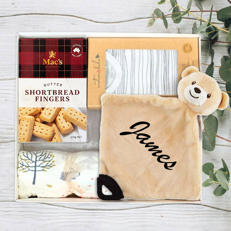 Gift set with Mac's shortbread fingers, a plush toy, and a personalized blanket on a wooden surface.