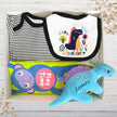 Baby gift box with bib, onesie, book, and plush blue dinosaur toy on a wooden surface.