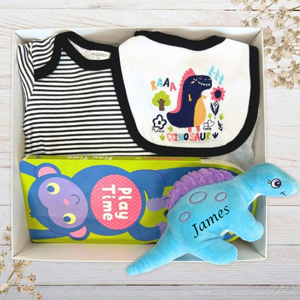 Baby gift box with bib, onesie, book, and plush blue dinosaur toy on a wooden surface.