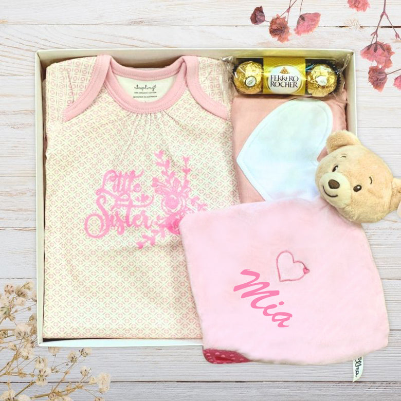 Baby gift set with little sister top, chocolates, and teddy bear in a box on a wooden surface.